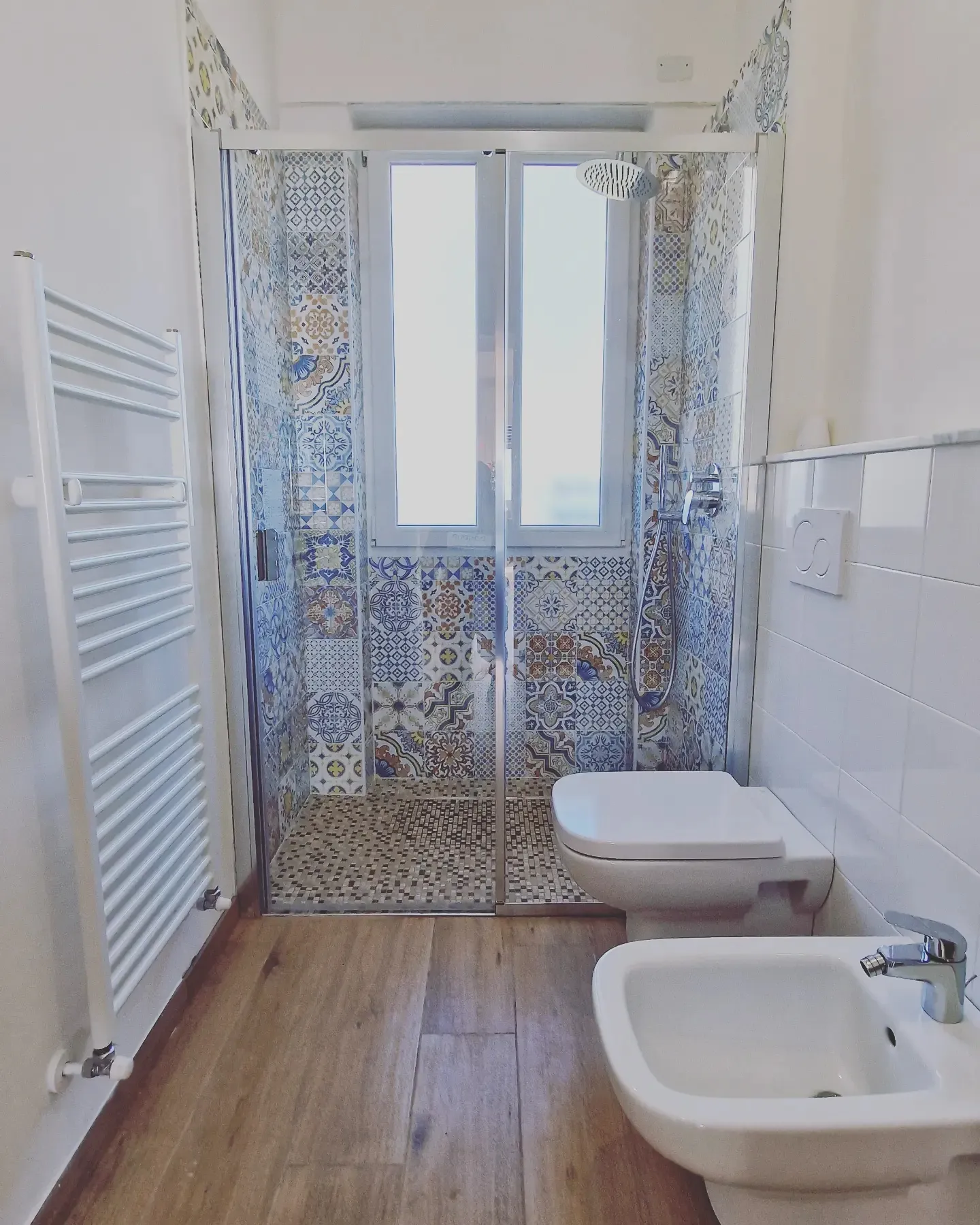 Bagno dopo la ristrutturazione: doccia in maioliche decorate sotto la finestra e pavimento effetto legno